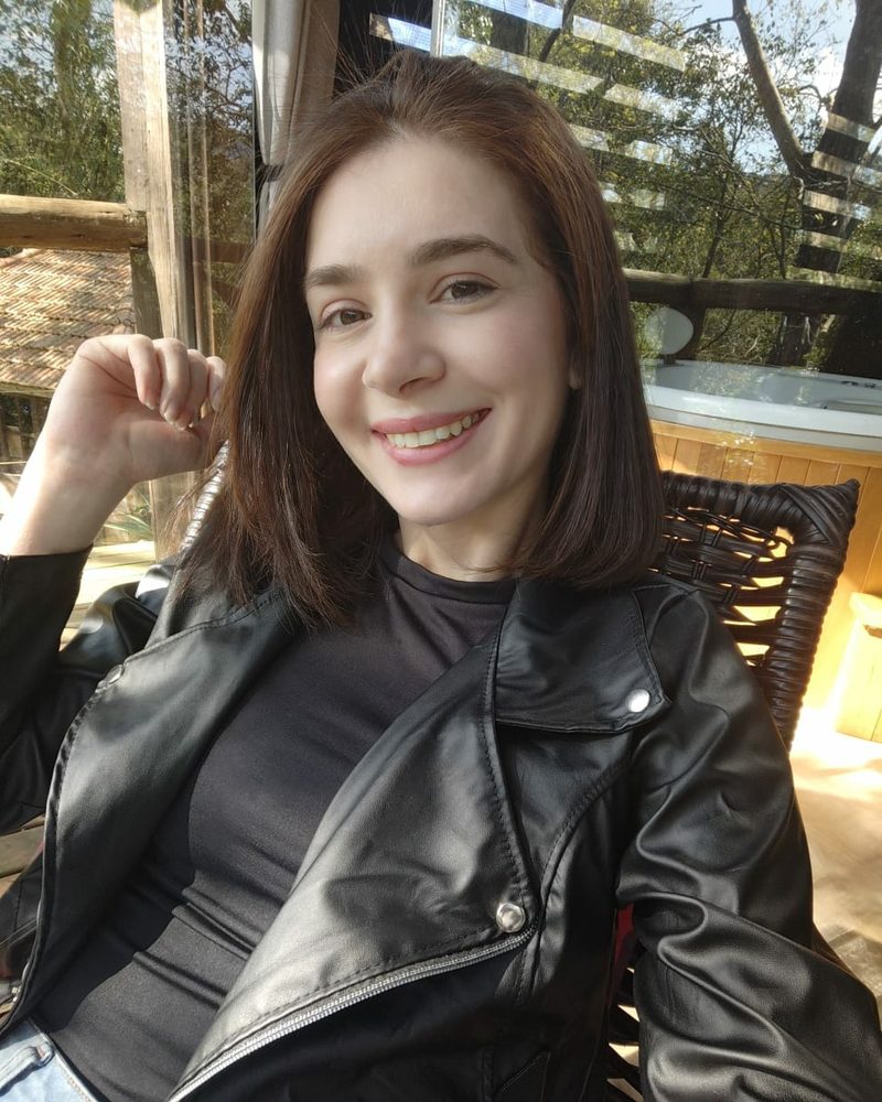 Simone sorrindo, de jaqueta de couro preta, sentada em uma varanda com árvores ao fundo